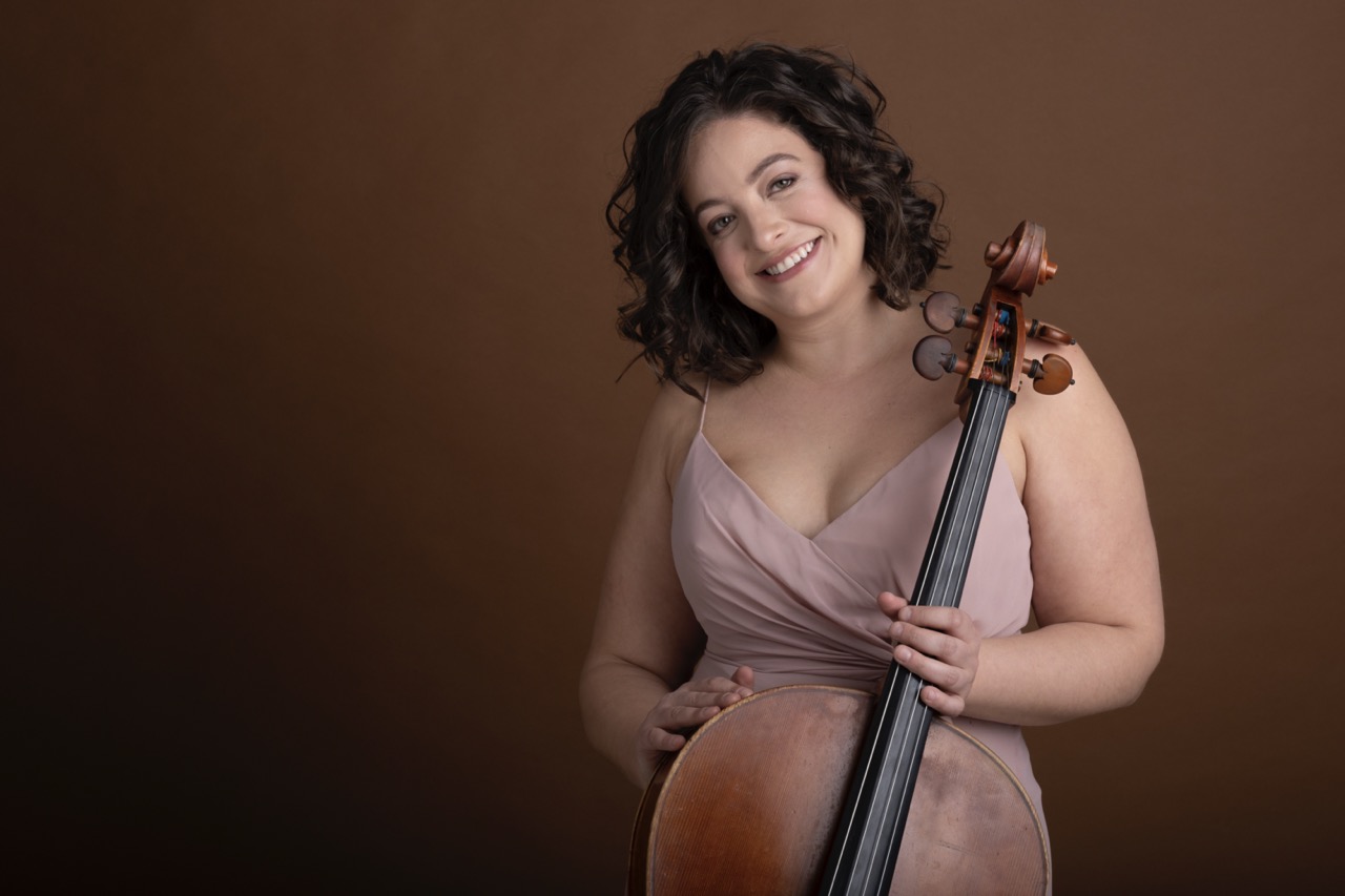 Gwen Krosnick stands on the right side of the frame in front of a brown background. She is holding her cello while wearing a pink dress.